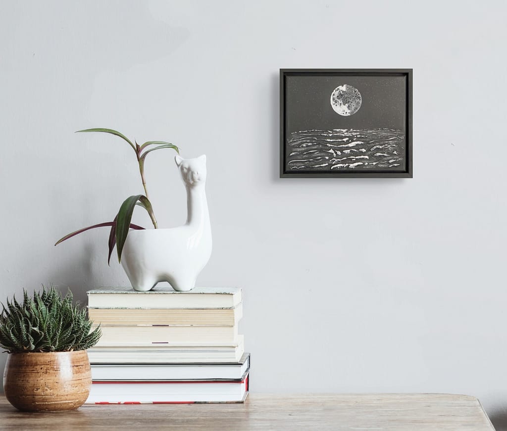 Small black and white moon and ocean painting hanging above a stack of books and a white llama planter